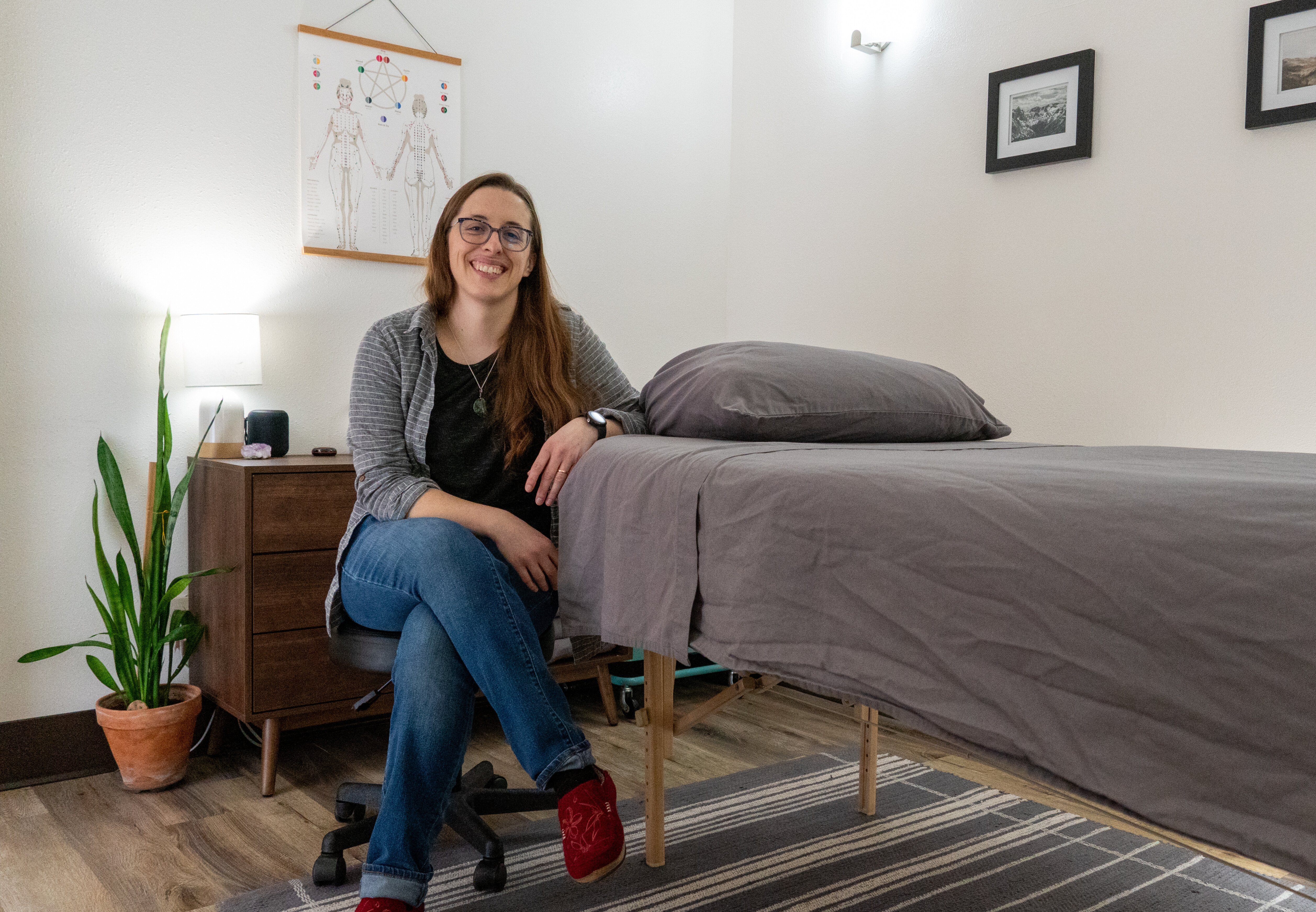 circular picture of Kate, a white woman, sitting in the treatment room in her clinic. Shes wearing jeans and a grey shirt, and has her arm resting on a massage table.